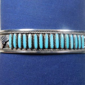 May include: Silver cuff bracelet with turquoise stones. The bracelet features a row of turquoise stones set in a silver band with leaf and rope detailing. The turquoise stones are a bright, vibrant blue color.