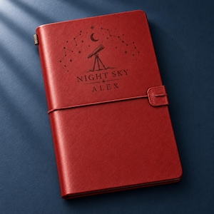 May include: A red leather-bound journal featuring an elastic closure strap and a metal pen loop. The cover is engraved with a telescope, a crescent moon, star constellations, and the text "NIGHT SKY" above the name "ALEX" in a clean font.
