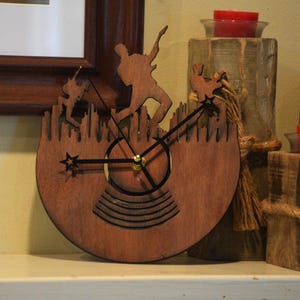 May include: A wooden clock with a brown finish, featuring a silhouette of a person playing guitar. The clock hands are black with a gold center. The clock is set against a background of wooden candle holders with red candles.