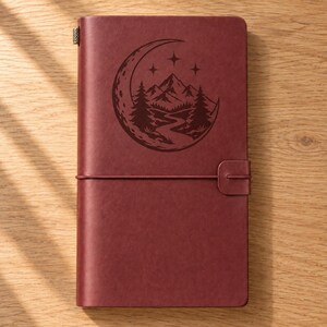 May include: A rectangular maroon notebook with a smooth cover featuring a dark engraved graphic of a crescent moon, stars, pine trees, and a winding path. The notebook is secured by a horizontal elastic band with a matching maroon tab closure.