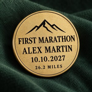 May include: A circular gold-toned metal coin featuring an engraved mountain silhouette at the top. The text reads FIRST MARATHON, ALEX MARTIN, 10.10.2027, and 26.2 MILES in black lettering. The coin has a smooth, matte finish and a raised border.