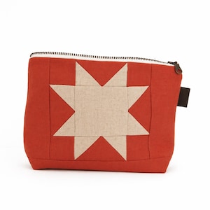 May include: A rectangular zippered pouch in a solid burnt orange fabric featuring a large, centered eight-pointed star applique in a light beige, textured fabric. The pouch includes a dark metal zipper closure along the top edge.