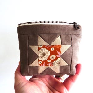 May include: A small, brown quilted pouch with a star design. The star is made of tan and orange fabric with a floral pattern. The pouch has a zipper closure and a tan lining.
