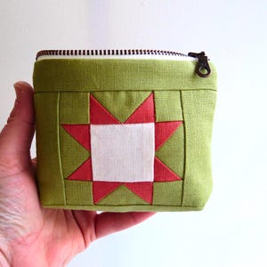 May include: A small, square, green fabric pouch with a star quilt block design. The star is red and white, and the pouch has a zipper closure. The pouch is held in a hand.