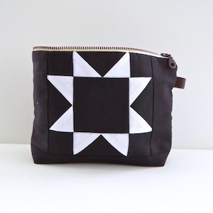 May include: A black zippered pouch featuring a central white star pattern made of geometric fabric pieces. The rectangular bag has a top metal zipper closure with a small brown leather pull tab attached to the side, creating a high-contrast design.