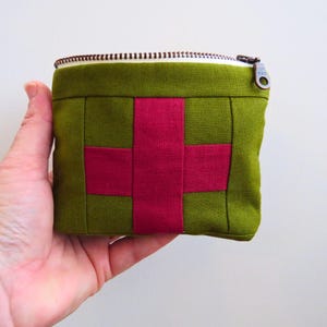 May include: A small olive green zippered pouch featuring a prominent magenta cross design on the front. The rectangular fabric accessory has a metallic zipper closure at the top and is constructed with visible seams forming a cross pattern.