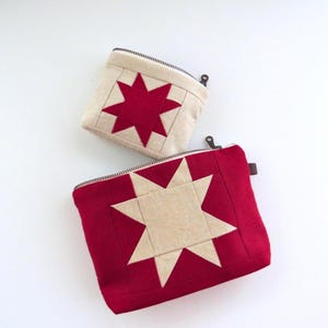 Puede incluir: Dos estuches con cremallera de tela. Uno es rojo con una estrella beige, el otro es beige con una estrella roja. Ambos tienen cremalleras negras. El diseño de la estrella es de estilo patchwork.