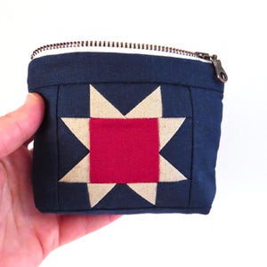 May include: A navy blue square pouch with a star quilt block design. The star is composed of a red center and tan points. The pouch has a white interior and a bronze zipper closure. The pouch is held in a hand.