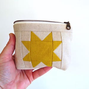 May include: A small, square, beige fabric pouch with a yellow starburst design. The pouch has a brown zipper and a bronze zipper pull. The starburst is centered on the front of the pouch.