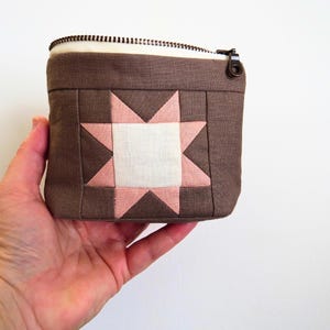 May include: A small brown fabric pouch featuring a central eight-pointed star design in light pink and white. The rectangular pouch has a top zipper closure with a dark metal pull. The exterior fabric has a visible woven texture throughout.