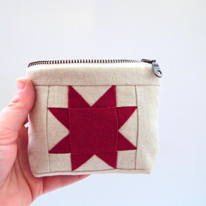 May include: A small, square, beige fabric pouch with a red star design on the front. The star is centered and made of several red triangles. The pouch has a zipper closure and a metal zipper pull.