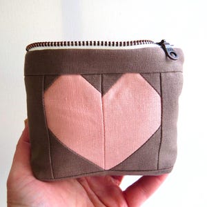May include: A small, square, brown and pink fabric pouch with a zipper closure. The front of the pouch features a large, pink heart design. The zipper is black with a brown pull. The pouch is held in a hand.