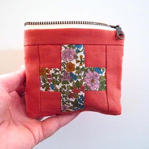 May include: A small, square, coral-colored fabric pouch featuring a central cross-shaped inlay of white fabric with a colorful floral pattern. The pouch is secured at the top with a metal zipper closure that has a dark-toned pull tab.