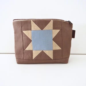 May include: A brown zippered pouch featuring a central star quilt block pattern. The star design is composed of a light blue square center surrounded by beige triangular points, all set against a solid brown fabric background with a dark zipper.