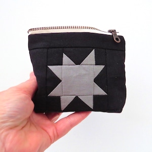 May include: A small black fabric pouch featuring a centered eight-pointed star design in light gray. The pouch has a top zipper closure with a metallic pull and a cream-colored interior lining visible along the top edge of the rectangular bag.