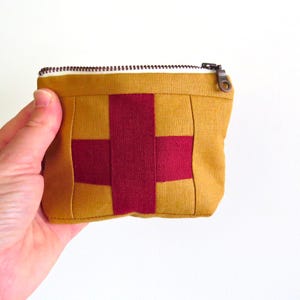 May include: A small, mustard-yellow and burgundy fabric pouch with a cross design. The pouch has a zip closure and is held in a hand against a white background. The cross is made of burgundy fabric, and the pouch is made of a woven material.