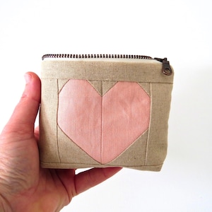 May include: A small, square, beige fabric pouch with a pink heart design on the front. The heart is made of pink fabric and is stitched onto the beige background. The pouch has a zipper closure with a bronze-colored pull.