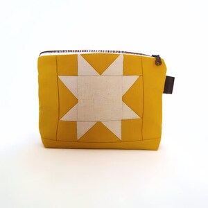 May include: A mustard yellow quilted pouch with a white starburst design. The pouch has a black zipper and a small brown leather pull tab. The design is reminiscent of a traditional quilt block.