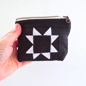 May include: A small, black fabric pouch with a white starburst design on the front. The pouch has a zipper closure and a bronze-colored zipper pull. The pouch is held in a person's hand.