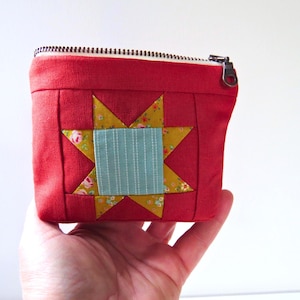 May include: A small, red quilted pouch with a star design. The star is composed of yellow triangles with floral patterns and a central light blue square. The pouch has a zipper closure.