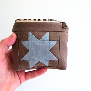 May include: A brown square zipped pouch featuring a light blue star design. The pouch has a bronze zip and pull. The star is centrally placed on the front of the pouch.