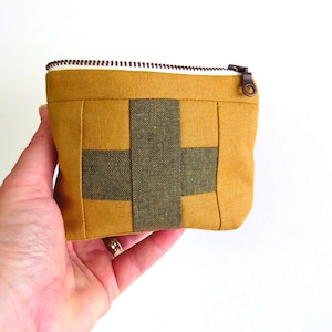 May include: A small, mustard yellow and olive green pouch with a zip closure. The square pouch has a woven pattern with a dark brown zip and a white lining. The pouch is held in a hand against a white backdrop.