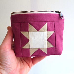May include: A small, square, quilted pouch in a deep magenta color. The front features a star design with a white center and tan points. The pouch has a white interior and a zipper closure with a metal pull.