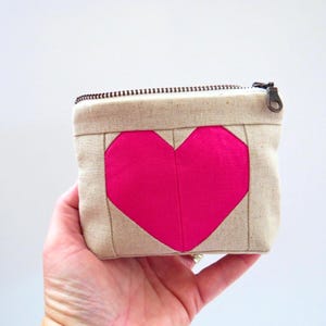May include: A small, square beige fabric pouch with a bright pink heart design on the front. The pouch has a zipper closure and a bronze-colored zipper pull. The heart is a geometric design.