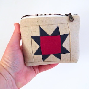 Puede incluir: Pequeño estuche de tela beige con diseño de estrella. La estrella está compuesta por un cuadrado rojo en el centro y puntas azul marino. El estuche tiene cierre de cremallera con tirador color bronce.