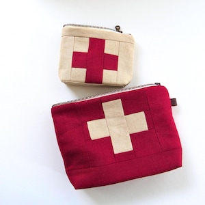 May include: Two zippered pouches with a cross design. One pouch is red with a tan cross, and the other is tan with a red cross. Both have a rectangular shape and a zipper closure.