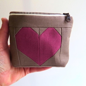 Puede incluir: Pequeño neceser cuadrado con exterior marrón y diseño de corazón magenta. El neceser tiene cierre de cremallera y se sostiene en una mano. El corazón es de tela y está centrado en la parte delantera.