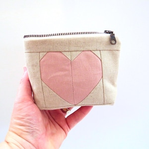 May include: A small, square, beige fabric pouch with a pink heart design on the front. The pouch has a black zipper and a bronze-colored zipper pull. The heart is made of a lighter pink fabric.