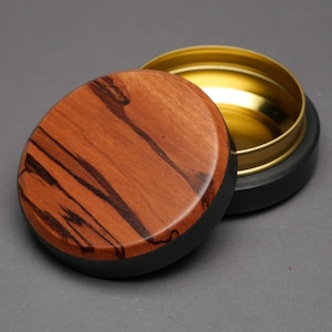 May include: A round wooden box with a lid. The lid is made of reddish-brown wood with dark brown streaks. The base of the box is black, and the interior is gold-colored. The lid is slightly ajar, revealing the interior.