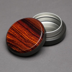 May include: An open, small, round container with a lid. The lid is made of wood with a striking pattern of orange and dark brown stripes. The container's body is silver, and the base is black.