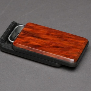 May include: A rectangular, black case with a hinged lid. The lid is made of wood with a reddish-brown, glossy finish. A silver-colored metal handle is visible inside the case. The case is partially open, revealing the interior.