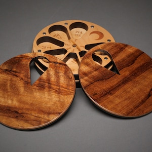 May include: Wooden time-telling device with a circular design. The device features a central rotating disc with cutouts for the hours and days of the week. Two wooden discs with handle-shaped cutouts are in the foreground. The wood has a rich, warm tone.