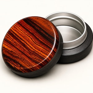 May include: A round container featuring a polished lid with a vibrant orange and dark brown wood grain pattern. The base of the container is matte black, and the interior is lined with a smooth, brushed silver-coloured metal insert.