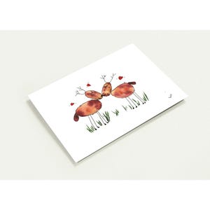 May include: A white card displays a watercolour illustration of four deer with brown bodies and thin legs. Each deer has small antlers, red hearts, and green grass. The card is angled, revealing a corner.