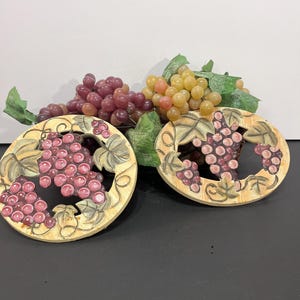 May include: Two round, cream-colored trivets featuring raised, hand-painted relief designs of purple and red grape clusters with green leaves. Behind the trivets are artificial bunches of red and yellow grapes with green foliage.