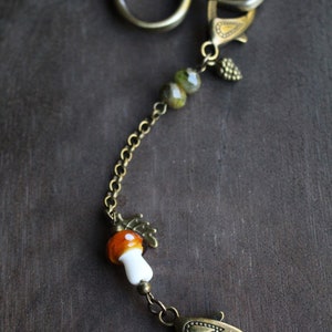 May include: A bronze-toned keychain with a whimsical mushroom charm. The charm features an orange and white mushroom, complemented by green beads and a small pinecone. The keychain has a lobster clasp and a chain.