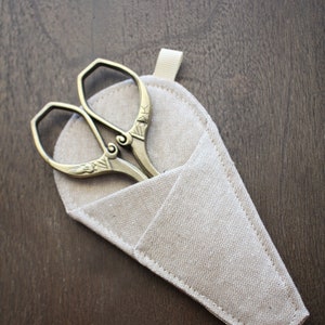 May include: A pair of antique gold scissors with ornate details, resting in a beige linen pouch with a white ribbon.