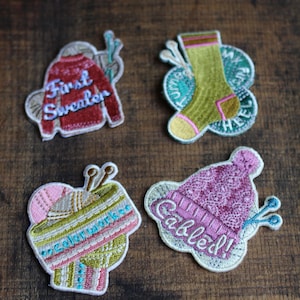 Könnte beinhalten: Vier gestickte Bügelbilder mit verschiedenen Strickmotiven. Die Patches befinden sich auf einem Holzgrund. Die Patches zeigen einen roten Pullover mit dem Text "First Sweater", eine grüne Socke mit dem Text "Heel to Toe", eine rosa Mütze mit dem Text "Cabled" und eine Wollknäuel mit dem Text "Colorwork".