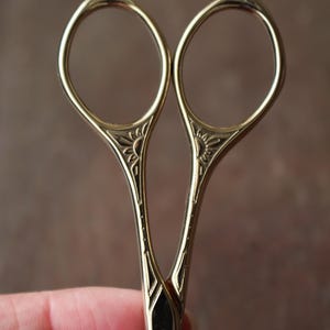 May include: Gold-colored metal embroidery scissors featuring symmetrical oval finger loops and a decorative engraved sunburst pattern at the base of the handles. The blades are joined by a central metal pivot screw, showcasing a polished finish.