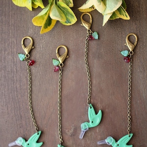 May include: Four metal chain charms featuring light green hummingbird-shaped pendants with clear plastic beaks. Each charm includes a bronze-toned lobster clasp, a small green leaf bead, and a tiny red faceted bead attached to the top of the chain.