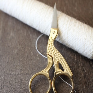 May include: Gold-colored metal embroidery scissors featuring a textured, bird-shaped handle design with a metallic finish and sharp, pointed silver-toned blades, positioned in front of a spool of off-white yarn with a single loose thread.