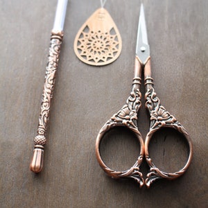 May include: A set of three copper-toned sewing tools: a pair of ornate scissors, a needle threader, and a small decorative metal piece with a circular design.