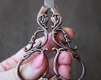 May include: Ornate copper-toned scissors with a decorative floral design. The scissors have a silver blade and are held in a hand.
