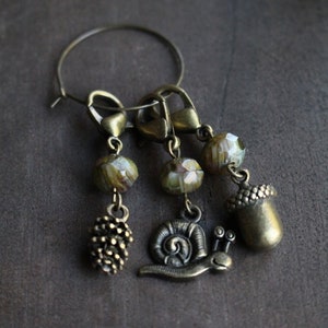 May include: A set of four stitch markers featuring a snail, acorn, pine cone, and three glass beads. The markers are attached to a silver ring.