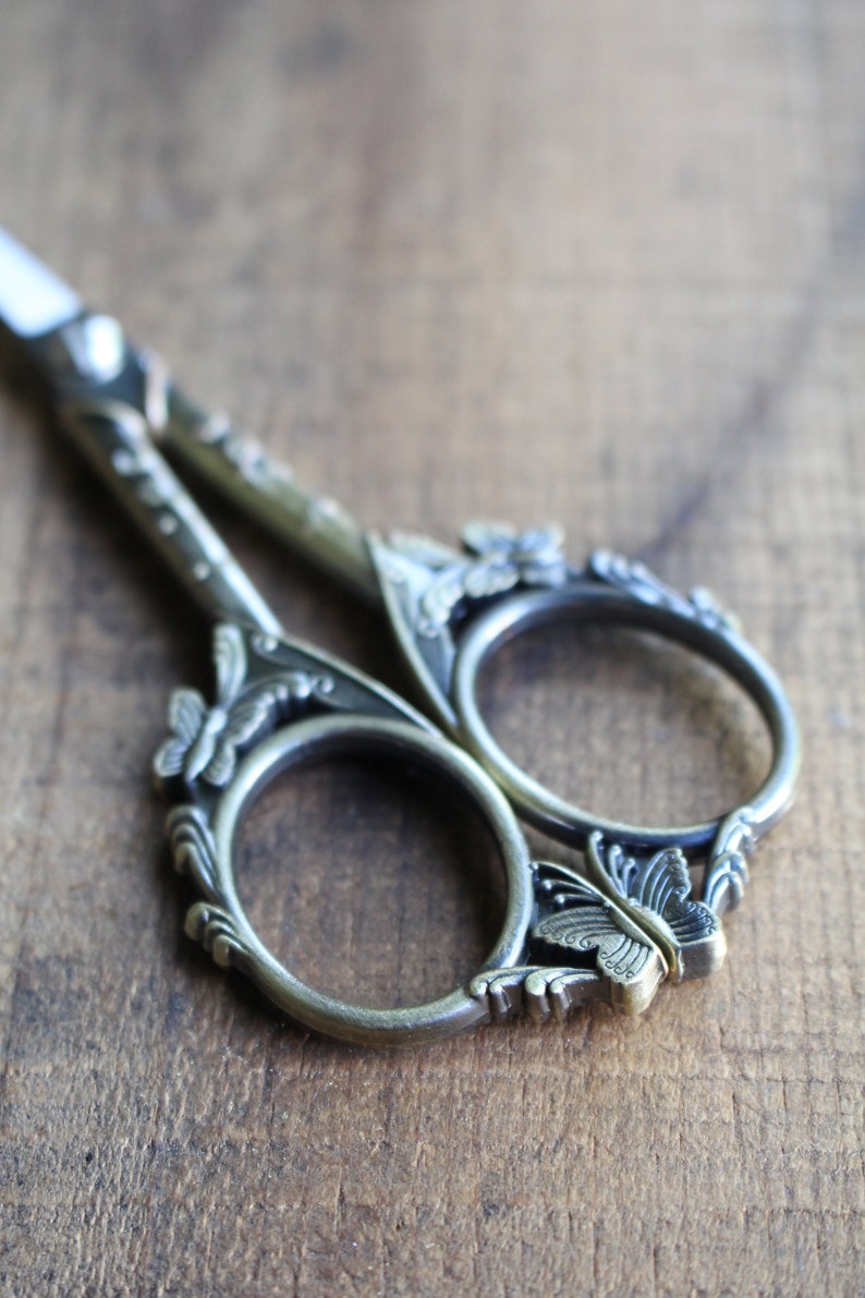 May include: Antique silver-toned scissors with ornate butterfly and floral designs. The scissors have a detailed, intricate design.