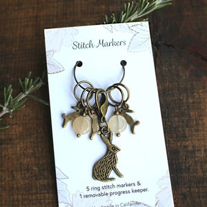 May include: A set of bronze-colored stitch markers and a removable progress keeper. The set includes five ring stitch markers and one progress keeper, each adorned with rabbit charms and beads. The card reads "Stitch Markers".